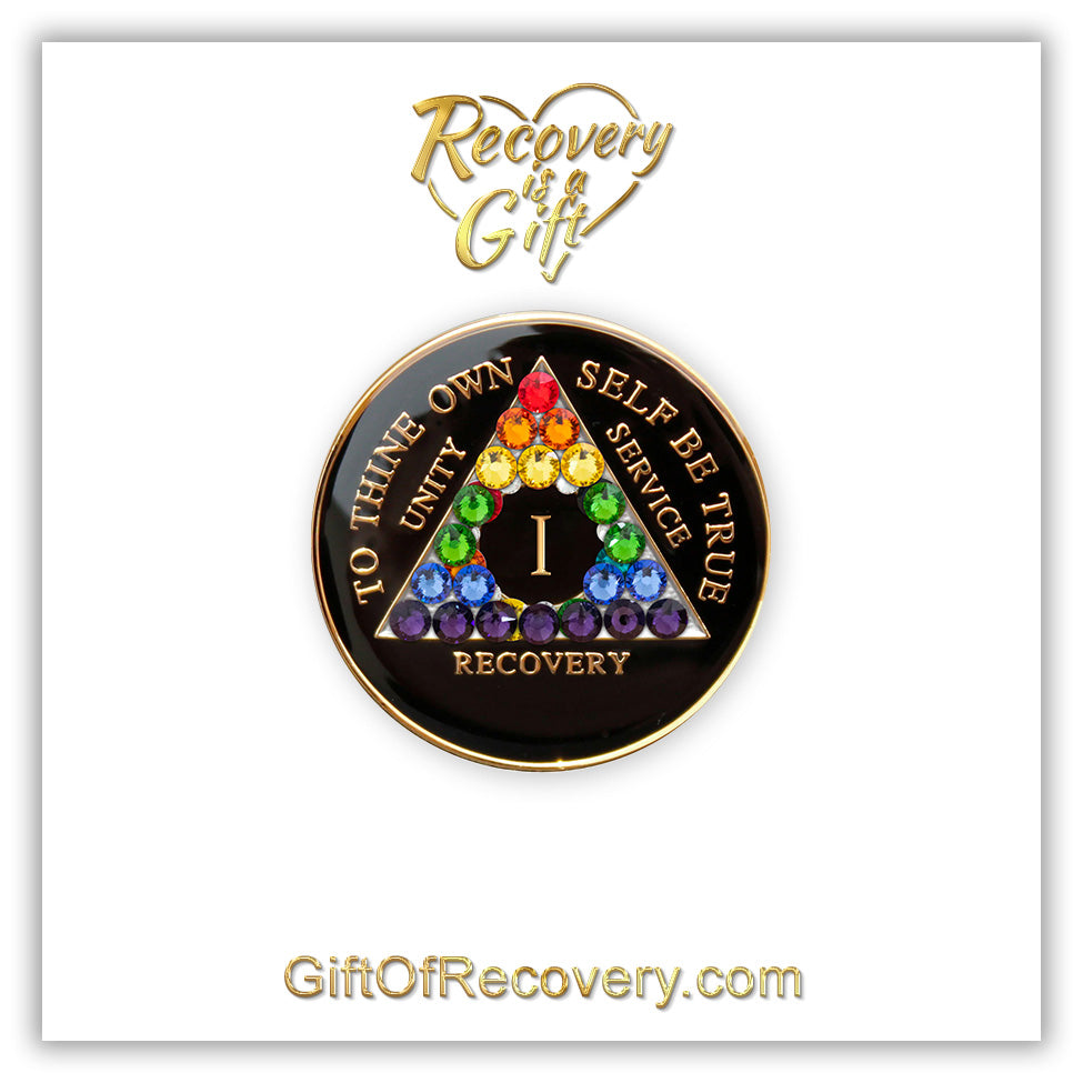1 year AA medallion with 21 rainbow genuine crystals to represent the LBGTQIA+ community, show your pride with this Black onyx resin sealed recovery medallion that has 14k gold plated brass lettering of to thine own self be true, unity, service, recovery, and the roman numeral, set on a white 3x3 card with recovery is a gift going through a heart in the color gold.