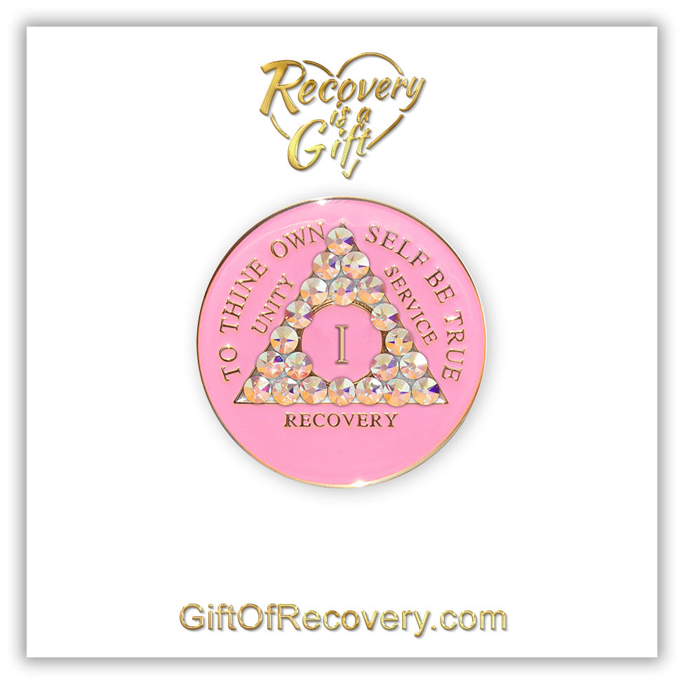 1 Year AA medallion princess pink with 21 Aurora Borealis CZ genuine crystals symbolizing beauty and change from working the program, AA slogan and 3 legacies are embossed with 14k gold-plated brass, sealed with resin for a lasting finish, seen on a white 3x3 card with recovery is a gift going through an open heart at the top and giftofrecovery.com at the bottom, both in the color gold.