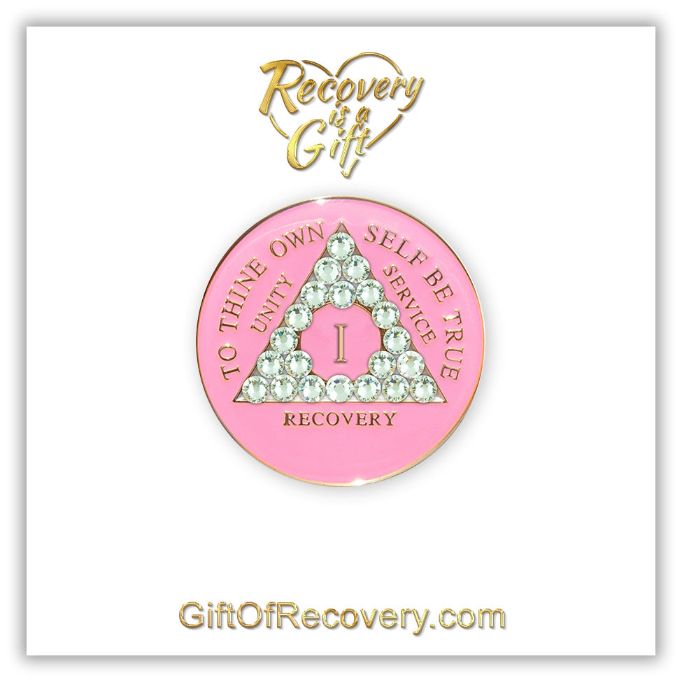 1 year AA medallion princess pink the triangle is adorned with 21 diamond CZ genuine crystals that represent transformation under pressure, the AA slogan and three legacies are embossed with 14k gold-plated brass, the recovery medallion is sealed with resin for a lasting finish, set on a white 3x3 card with recovery is a gift going through an open heart at the top and giftofrecovery.com on the bottom, both in the color gold.