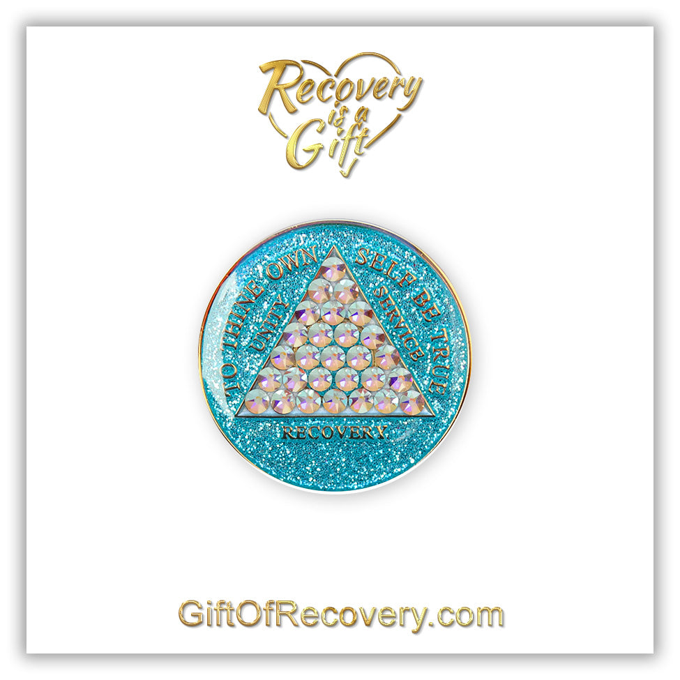 AA medallion Aquamarine glitter with 28 diamond CZ genuine crystals that form the triangle entirely, this is the timeless medallion, shine on with this beauty, we all have one day at a time, the AA slogan and three legacies are embossed with 14k gold-plated brass and sealed with resin for a glossy finish that will last and is scratch proof, pictured on a white 3x3 card with recovery is a gift going through an open heart at the top and giftofrecovery.com at the bottom, both in the color gold.