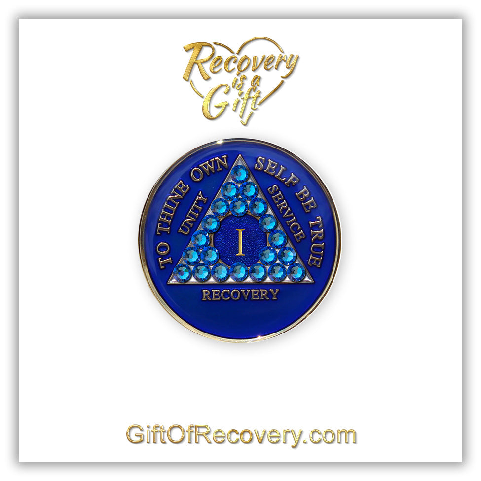 1 year AA medallion Big Book blue adorned with 21 genuine blue CZ crystals symbolizing the importance of our Big Book, to thine own self be true, unity, service, recovery are embossed with 14k gold-plated brass and sealed with resin for a glossy finish that is scratch proof and will last, seen on a white 3x3 card with recovery is a gift going through an open heart at the top and giftofrecovery.com on the bottom, both in the color gold.