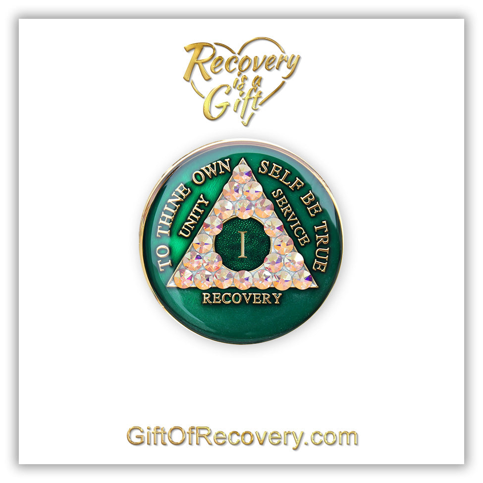 1 year AA medallion Emerald green with 21 genuine Aurora Borealis CZ crystals that make the triangle, to symbolize new beginnings and to dance in the sunlight once again, AA slogan and three legacies are embossed with 14k gold-plated brass and sealed for a glossy finish that will last, pictured on a white 3x3 card with recovery is a gift going through an open heart at the top and giftofrecovery.com at the bottom, both in the color gold.