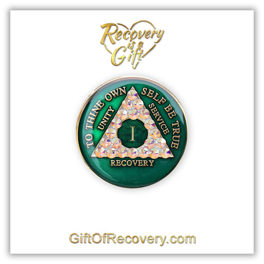 1 year AA medallion Emerald green with 21 genuine Aurora Borealis CZ crystals that make the triangle, to symbolize new beginnings and to dance in the sunlight once again, AA slogan and three legacies are embossed with 14k gold-plated brass and sealed for a glossy finish that will last, pictured on a white 3x3 card with recovery is a gift going through an open heart at the top and giftofrecovery.com at the bottom, both in the color gold.