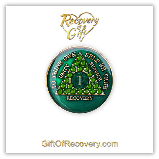 1 year AA medallion Emerald green with 21 genuine green crystals that make the triangle, to represent luck and prosperity in the program, AA slogan and three legacies are embossed with 14k gold-plated brass and sealed for a glossy finish that will last, pictured on a white 3x3 card with recovery is a gift going through an open heart at the top and giftofrecovery.com at the bottom, both in the color gold.