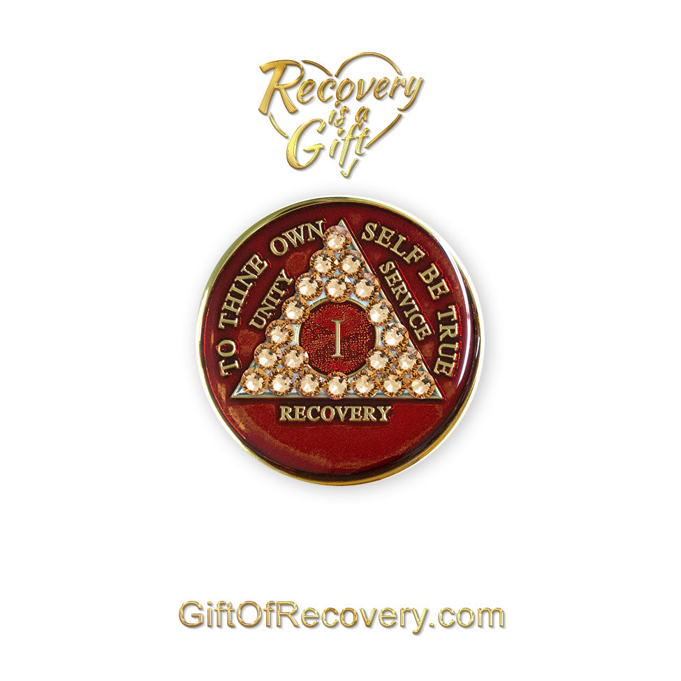 1 year ruby red AA medallion adorned with 21 genuine gold CZ crystals symbolizing strength and elegance in recovery, to thine own self be true, unity, service, recovery are embossed with 14k gold-plated brass and sealed with resin for a glossy finish that is scratch proof and will last, seen on a white 3x3 card with recovery is a gift going through an open heart at the top and giftofrecovery.com on the bottom, both in the color gold. 