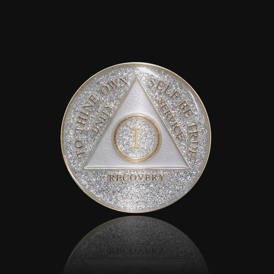 1 year AA medallion Glitter silver, always shine on in sobriety, triangle is pearl white, to thine own self be true, unity, service, recovery, and roman numeral are embossed with 14k gold-plated brass, the recovery medallion is sealed with resin for a glossy finish that will last and is scratch free, pictured on a white 3x3 card with recovery is a gift going through an open heart at the top and giftofrecovery.com on the bottom, both in the color gold.